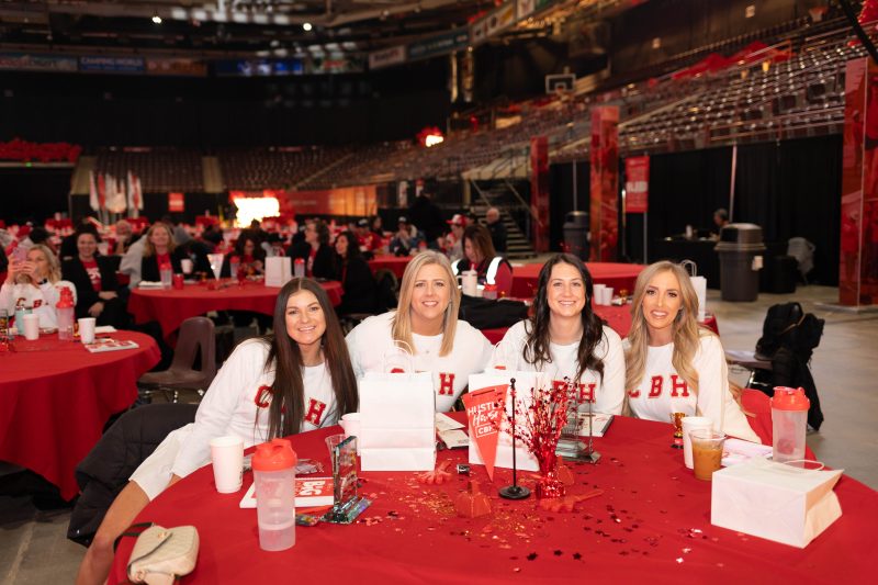 Four smiling women in matching white CBH Homes shirts sit together at a red-decorated table during the 2025 CBH Homes Company Kickoff event. The event is held in a large indoor arena, with employees gathered at tables covered in red decorations, CBH-branded items, and event materials. Attendees in the background engage in conversation, capturing the excitement and team spirit of the annual company celebration. This vibrant corporate event highlights CBH Homes' commitment to teamwork, motivation, and growth in the homebuilding industry.